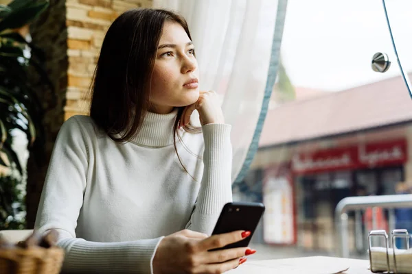 Pensive young brunette girl sitting in cafe, holding smartphone and looking out the window — Stock Photo, Image