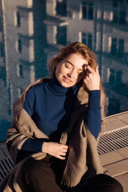 Mavi kazaklı sarışın kadın havuz kenarında oturur genç gülümseyen, güneş ve sıcaklık, gülüyor sahiptir