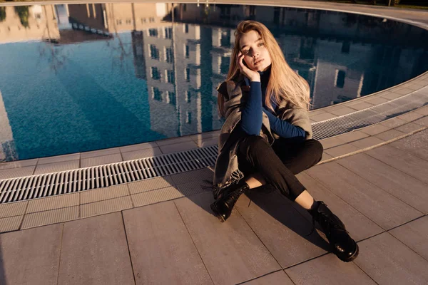 Elegante ragazza bionda seduta vicino alla piscina blu, sulle spalle calda sciarpa, godendo il sole — Foto Stock