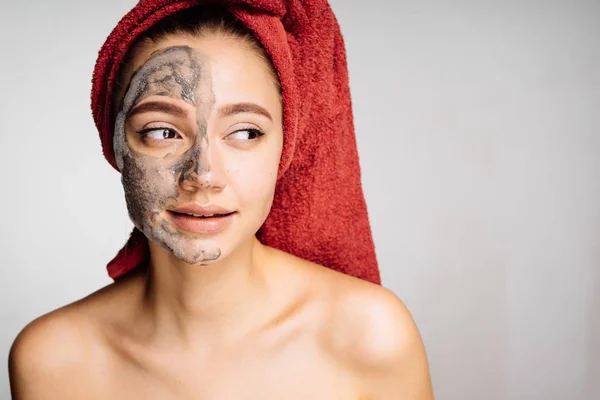 Hermosa joven con una toalla roja en la cabeza se aplica una máscara de arcilla a la mitad de la cara — Foto de Stock