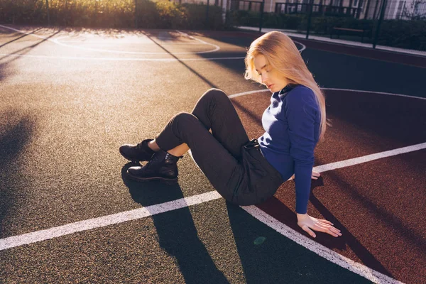 Menina modelo loira elegante em camisola azul senta-se no parque infantil posando em raios de sol da manhã — Fotografia de Stock