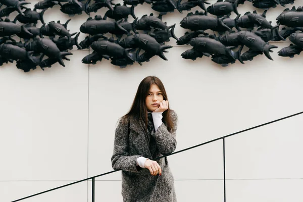 Réfléchie jeune fille aux cheveux bruns dans un manteau gris se dresse sur le fond d'un mur inhabituel — Photo