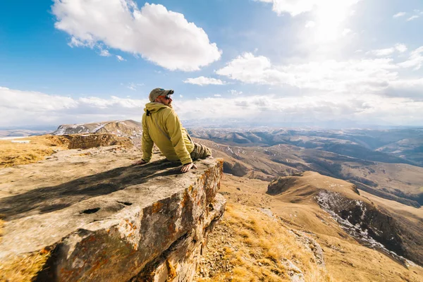 Ein männlicher Reisender in Jacke sitzt am Rande einer Klippe und genießt die Natur der Berge und die saubere Luft — Stockfoto