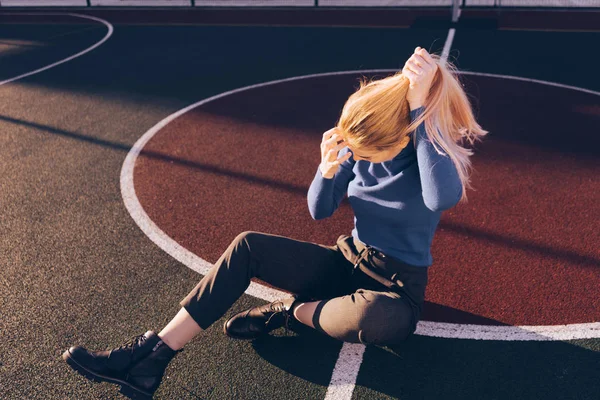Stijlvolle jong meisje met lang blond haar in een blauwe trui zit op het sportveld — Stockfoto