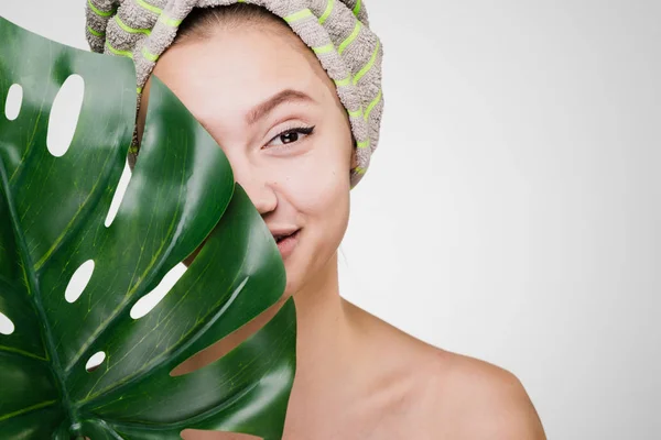 Leende ung flicka med ren hud innehar ett grönt blad, på huvudet en handduk, ett dagspa — Stockfoto