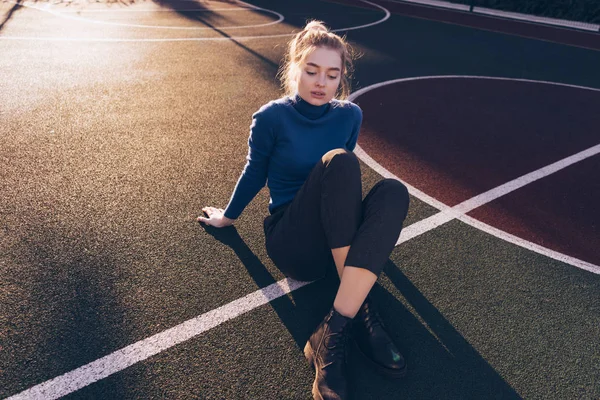 Stijlvolle jonge blonde model meisje in blauwe trui poseren op sportveld in de ochtendzon, Streetstyle en mode — Stockfoto