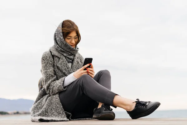 A stylish dark-haired young girl with glasses, a headscarf on her head, sits outdoors, writes a message on the smartphone — Stock Photo, Image