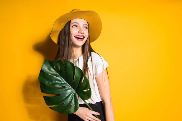 Bella ridendo giovane ragazza che tiene una foglia verde, sulla testa un grande cappello alla moda — Foto Stock