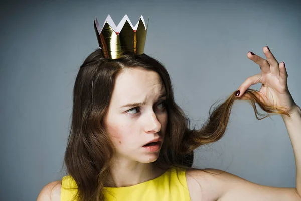 Joven insatisfecha sosteniendo un rizo de pelo, sobre su cabeza una corona dorada — Foto de Stock
