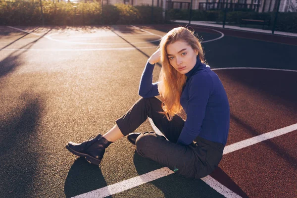 Стильна красива дівчина модель блондинка в синьому светрі сидить на спортивному полі на землі і позує — стокове фото