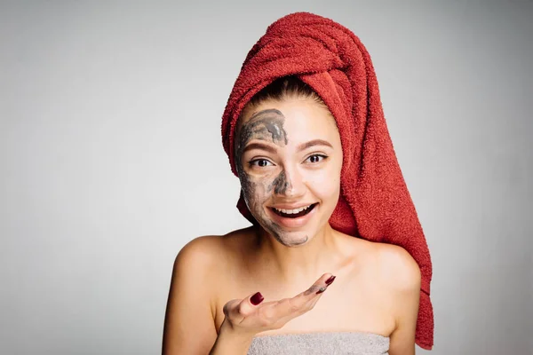 Happy laughing young girl with a red towel on her head puts a clay mask on her face — Stock Photo, Image