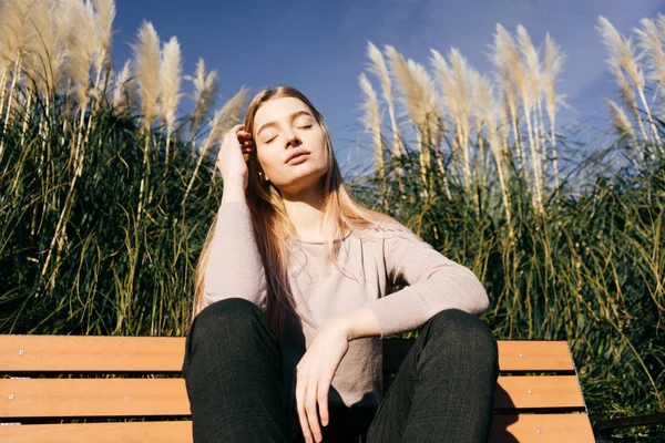 Mooie stijlvolle jong blond meisje zittend op een bankje buiten in de zon — Stockfoto