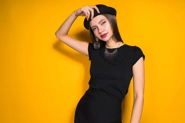 Confident beautiful girl in black dress and fashionable hat posing on orange background, smiling — Stock Photo, Image