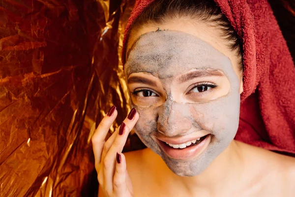 Ευτυχισμένος χαμογελαστό κορίτσι με μια κόκκινη πετσέτα στο κεφάλι της βάζει μια χρήσιμη μάσκα στο πρόσωπό της, ημερήσιο σπα — Φωτογραφία Αρχείου