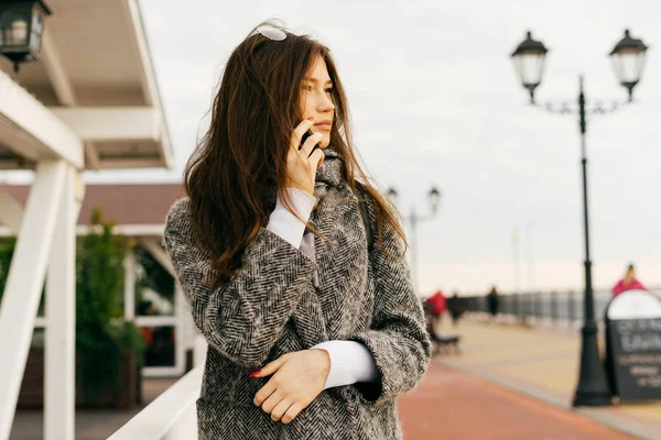 Élégante jeune fille brune en manteau gris parlant au téléphone, à l'extérieur, journée nuageuse, mode de rue — Photo