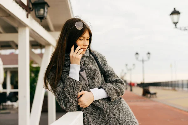 Стильна молода брюнетка в сірому пальто говорить по телефону, на відкритому повітрі — стокове фото