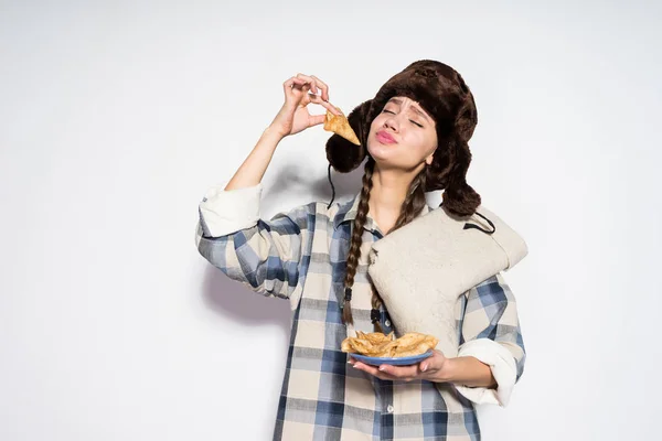 Una giovane e divertente ragazza russa mangia frittelle calde deliziose, cappello di pelliccia sulla testa, celebra Shrovetide — Foto Stock