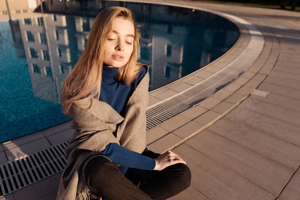 Ragazza in maglione blu siede a bordo piscina, gode del sole — Foto Stock