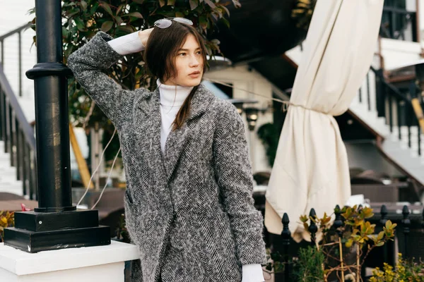 Chica joven de pelo oscuro con estilo en un abrigo gris camina por las calles de la ciudad, en sus gafas de cabeza — Foto de Stock