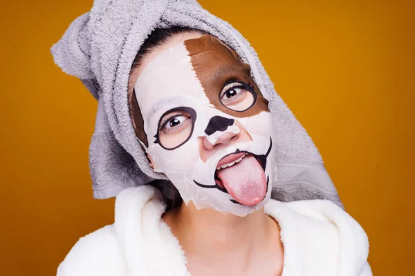 Ung flicka med en handduk på huvudet, på hennes ansikte en mask med en bild av en munkorg av en hund, visar en tunga — Stockfoto