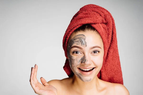 Una linda joven con una toalla roja en la cabeza se puso una máscara de barro en la mitad de su cara, sonriendo —  Fotos de Stock