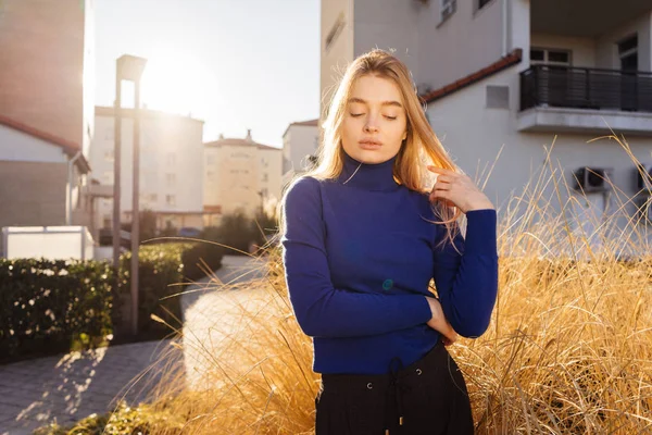 Jovem menina loira bonito em uma camisola azul andando pelas ruas da cidade, apreciando o clima quente e sol — Fotografia de Stock
