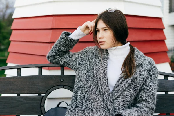 Confiante fille aux cheveux bruns en manteau gris s'assoit sur un banc à l'extérieur, style de rue — Photo