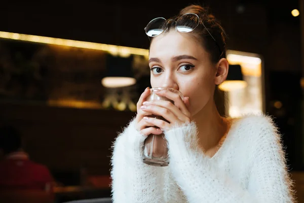 Милая юная студентка в белом свитере пьет капучино в кафе — стоковое фото