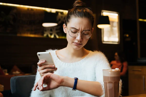 Business young girl freelancer with glasses holds smartphone and looks at clock — Stock Photo, Image