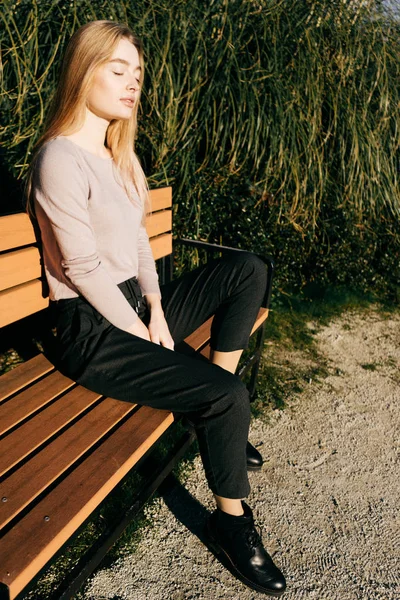 Élégante jeune fille blonde assise sur un banc au soleil, à l'extérieur, les yeux fermés — Photo
