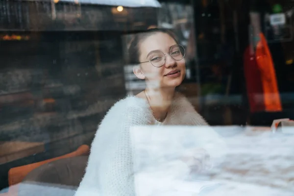 Красива молода дівчина-фрілансер в білому светрі і окулярах сидить в кафе і посміхається — стокове фото