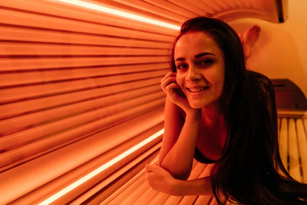 Una joven sonriente yace en una cama de bronceado bajo los rayos ultravioleta, tomando el sol, quiere una piel bronceada — Foto de Stock