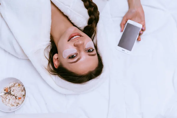 Chica en una túnica blanca se acuesta en la cama, temprano en la mañana, junto con parches de ojos — Foto de Stock