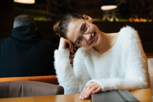 Мила молода дівчина в окулярах і біла куртка сидить в кафе після роботи і посміхається — стокове фото