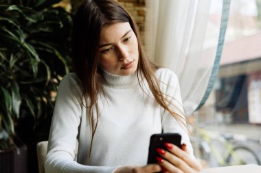 dalgın genç kız beyaz kazaklı bir kafede oturur ve onu smartphone içinde görünüyor