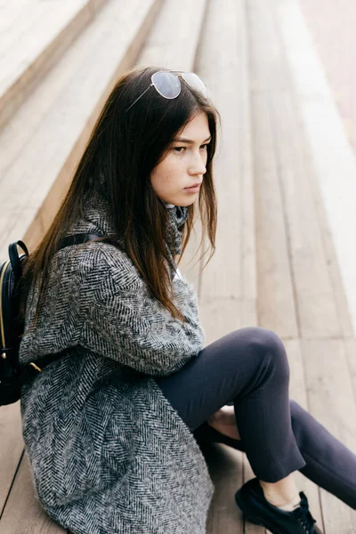 Young dark-haired girl in a fashionable gray coat sits on the steps in the streets of the city — Stock Photo, Image