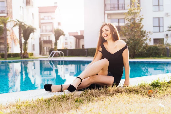 Смеющаяся шикарная девушка в черном платье и туфлях сидит у синего бассейна, наслаждается богатой жизнью и отдыхом — стоковое фото