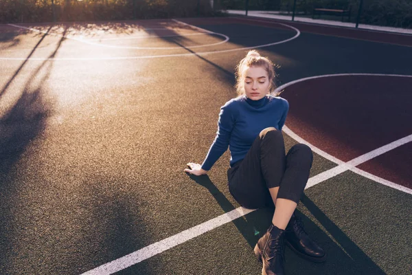 Jovem loira modelo menina em azul camisola da moda senta-se no chão em um campo de esportes posando — Fotografia de Stock