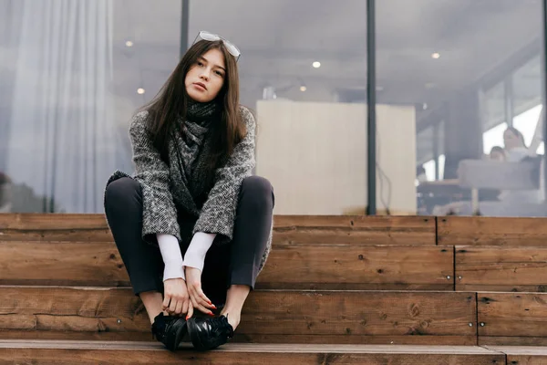 Stylish dark-haired young girl in trendy gray coat sits on steps, posing — Stock Photo, Image