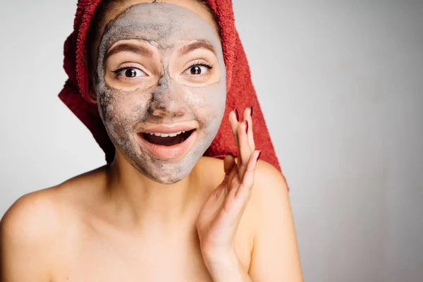 Happy νεαρό κορίτσι με μια πετσέτα στο κεφάλι της ένα χρήσιμο πήλινο προσωπείο εφαρμόζεται στο πρόσωπό της — Φωτογραφία Αρχείου