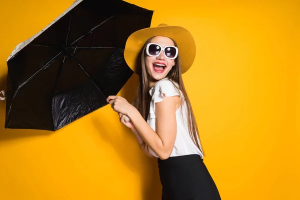 在黄色背景下, 戴着雨伞的太阳镜和帽子的快乐年轻女孩 — 图库照片