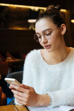 güzel genç kız freelancer gözlük ve beyaz bir ceket ile bir akıllı telefon tutar, bir kafede oturuyor