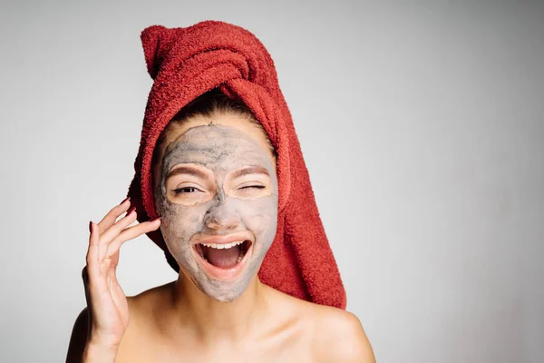 Niña feliz con una toalla roja en la cabeza se aplica una máscara de arcilla útil en la cara, se ríe —  Fotos de Stock