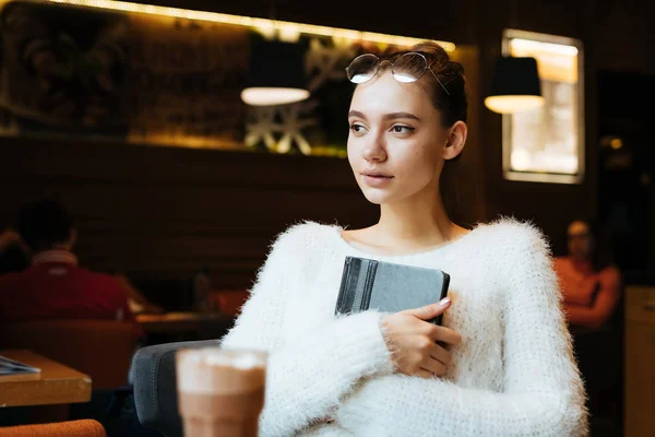 Joven hermosa freelancer chica con gafas y un suéter blanco se sienta en un café, sostiene un cuaderno para el trabajo —  Fotos de Stock