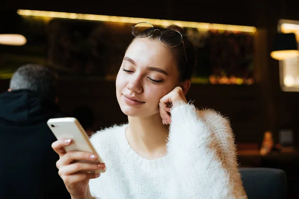 Cute young girl student in a white jacket is sitting in a cafe, looking into her smartphone — Stock Photo, Image