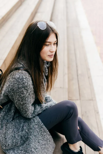 Милая темноволосая девушка в сером пальто сидит на ступеньках под открытым небом, в уличном стиле и моде — стоковое фото