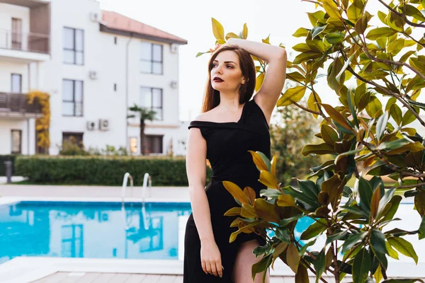 Luxurious attractive woman in a black dress posing in the garden by the blue pool, enjoying a rich life — Stock Photo, Image