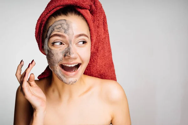 En skrattande ung flicka med en röd handduk på huvudet gäller en användbar lermask på läpparna, ett dagspa — Stockfoto