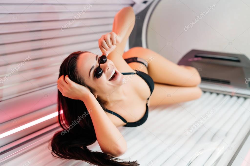 attractive happy girl lies in a horizontal tanning bed, wearing safety glasses, sunbathes and smiles