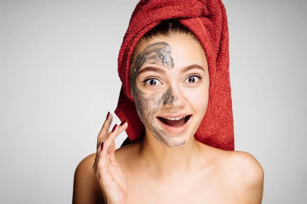 Happy young girl with a red towel on her head applies a useful clay mask on the face — Stock Photo, Image